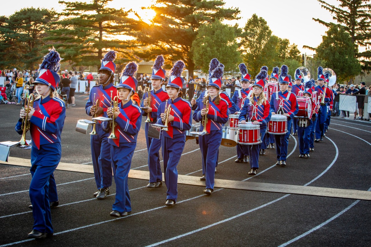 The marching band, in uniform at a football game, marching to the field to play their halftime show.